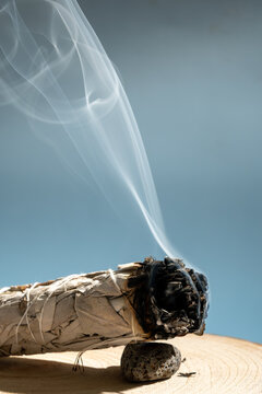 Burning white sage smudge stick releasing soft smoke, placed on a natural wooden surface against a calm blue background