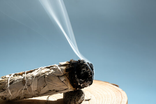 white sage smudge stick burning on wooden surface emitting gentle smoke against clear blue sky