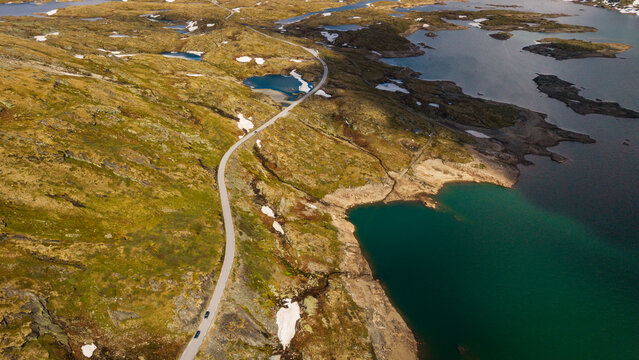 Aerial view of Sognefjellet mountain road winding through the rugged landscape with glacial lakes, rocky terrain, and patches of snow in Lom Municipality, Innlandet, Norway.
