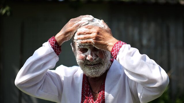 Senior man wearing in ukrainian vyshyvanka lathering his hair with soap at summer garden. The scene captures care with a sense of peace and natural simplicity. Concept of tradition and daily ritual
