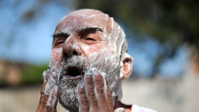 Elderly man wearing in traditional embroidered shirt washing his gray beard fully covered in soap at summer garden. Scene captures care with sense of peace and natural simplicity. Daily ritual concept