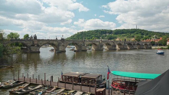 Historic Charles Bridge spans the Vltava River in Prague. Wooden boats moor at the river pier in the foreground. Green Petrin Hill rises under a cloudy summer sky for European travel concepts.