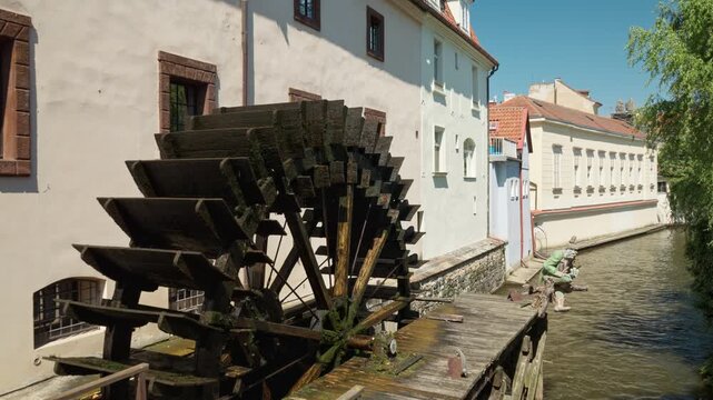 Historic wooden water wheel turns on Certovka canal in Prague. Grand Priory Mill mechanism rotates near traditional buildings. Ideal European travel destination for summer city tour.
