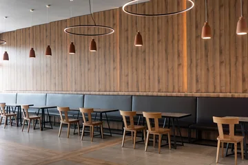 Fotobehang Horeca Interior view of an empty modern restaurant featuring stylish wooden chairs along a long black leather banquette with contemporary circle pendant lights hanging against a warm textured wood wall.   © Christos
