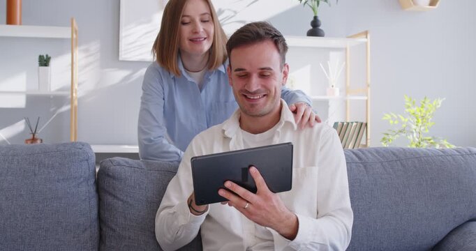Couple using tablet browsing internet for social media and online shopping. On sofa at home, they browse a bright room, compare items, and prepare payment with a secure digital app. 