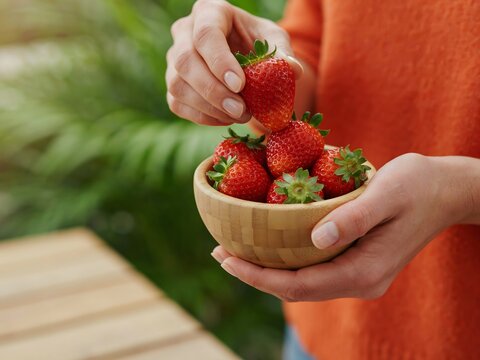Person holding a small wooden bowl filled with fresh ripe red strawberries outdoors