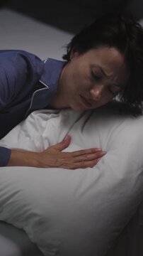 A woman in her 30s lies on a bed in a dark room trying to fall asleep while wearing blue pajamas and holding a pillow close.