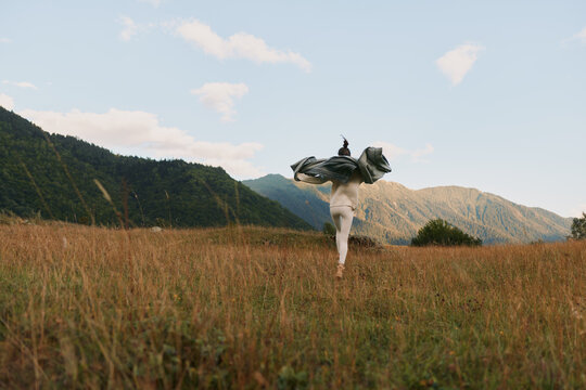 Woman lifestyle outdoors running through tall grass field with flowing jacket mountains background clear sky natural light freedom and movement