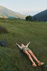 Obraz premium A young adult woman lies on a grassy field with arms outstretched while distant mountains rise, conveying a moment of relaxation, freedom, and carefree outdoor living.