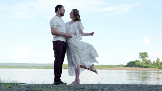 Standing lakeside, woman lifts dress hem while man supports her waist beside calm water under soft daylight, creating elegant outdoor fashion scene with relaxed pose and quiet shoreline mood