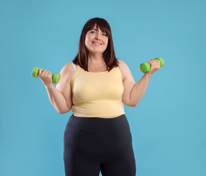 Overweight woman exercising with dumbbells on light blue background. Sports equipment