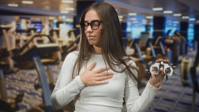 Woman optician wearing black glasses holds an eyeglass trial frame in one hand, other hand on chest with eyes closed inside a building testing lenses; thoughtful focus.