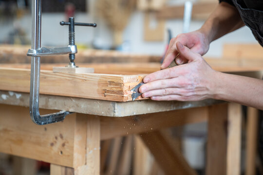 Professional handicraftsman processes edges of antique wooden window frame with chisel instrument, master carefully cutting and smoothing surface of frame detail fixated at workbench with clamp tool.