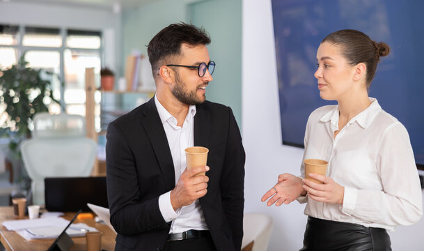 Man and a woman work in an office, they stand near a table and drink hot coffee during a work break
