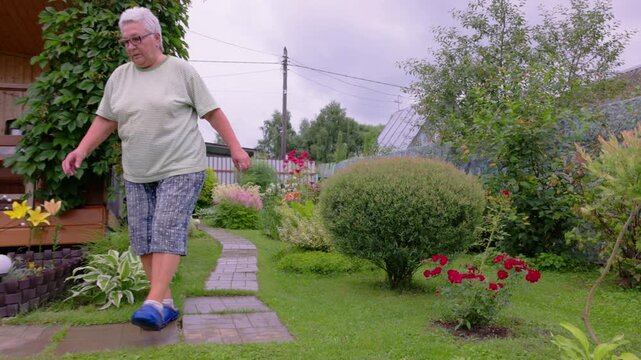Senior woman walking towards her country house in the beautiful blooming garden. Elderly female gardener walking down the path past beautiful flowers on her backyard. 