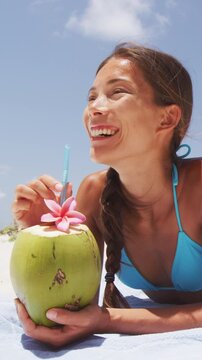 Happy beach bikini woman relaxing drinking fresh coconut water lying down sunbathing on fun Caribbean vacation tanning under the tropical sun. Asian model enjoying suntan.