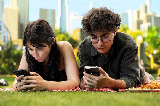 Young couple lie side by side on a picnic blanket and ignore each other while doomscrolling on smartphones, showing phubbing, poor communication, digital distraction, and relationship disconnection.