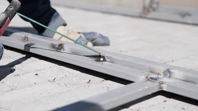 Worker welds steel outdoors with intense blue white arc, small sparks, smoke haze, gloved hand guiding rod along seam, low angle diagonal view over crossing metal bars, soft daylight, muted gray
