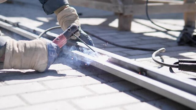 Close view shows gloved worker welding steel bars on rooftop surface, with bright blue arc at joint, orange sparks, drifting smoke, cable, torch, and softly blurred support structure under hard