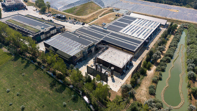 Aerial view of an integrated waste management facility with solar panels on rooftops, industrial buildings, and an adjacent pond in Cascina Maggiore, Lombardy, Italy.