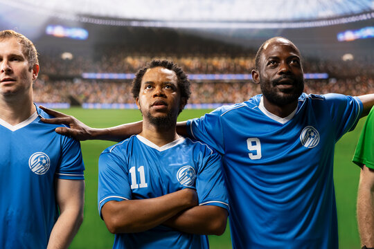 Diverse soccer players and goalkeeper stand together in a pre-match anthem lineup on the pitch, showing team unity with the stadium crowd blurred behind them during the opening ceremony.