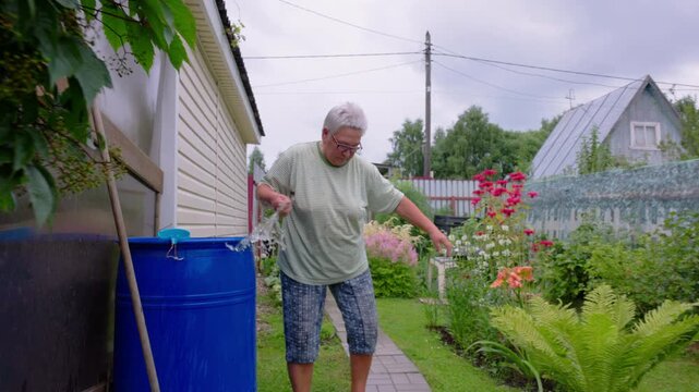 Senior woman washing hands in a blue barrel with rainwater in the garden after planting flowers.
