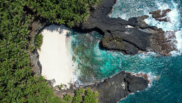 Praia Escada on Ilh&eacute;u das Rolas, S&atilde;o Tom&eacute; and Pr&iacute;ncipe. Aerial view of a secluded white-sand cove surrounded by volcanic rock formations, coconut palm forest, and crystal-clear turquoise waters