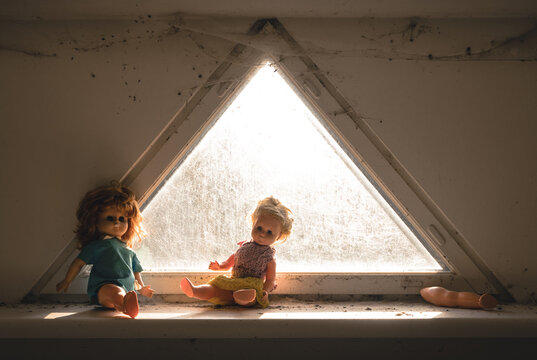 View of two dolls sitting on a dusty windowsill in front of a triangular window with cobwebs and a detached doll leg in Tallinn, Harju County, Estonia.