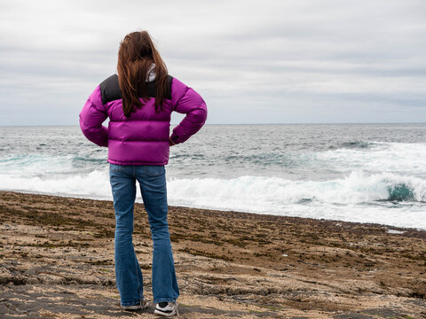 Girl in pink jacket stands facing ocean waves. She wears jeans and sneakers on rocky shore. Sky overcast, sea water crashes nearby. Scene shows solitude and nature power. Travel Ireland.