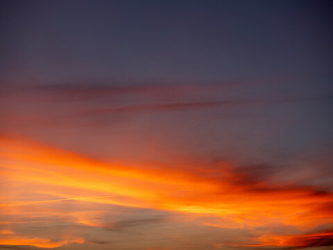 Dramatic sky at sunset with streaky clouds. Warm orange and yellow hues glow near horizon. Darker clouds contrast with soft light. Sky texture shows natural movement