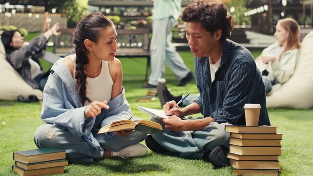 Students sit on the grass beside stacked books and take notes together, using coursework and textbooks as focused peers discuss exam preparation during an outdoor campus study session.