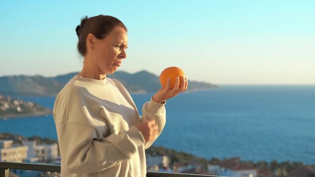 Woman suffering allergic reaction holding orange on balcony. Adult woman standing on a balcony with a sea view, experiencing an allergic reaction, itching her neck and face after smelling an orange
