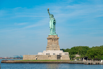 Statue of Liberty overlooks New York Harbor on Independence Day. Wide daylight view frames Liberty Island, stone pedestal, green trees and blue water beneath a clear sky. © white78