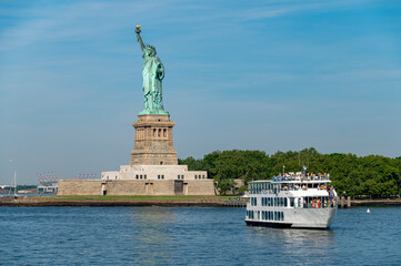 New York, USA, 4 Jule 2026 : Tour boat crosses New York Harbor beside Statue of Liberty. Passengers stand on three decks of a white vessel with Liberty Island trees and calm water behind. © white78