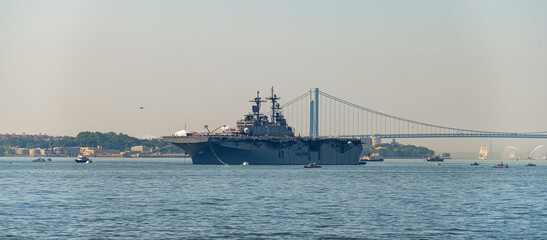 USS Kearsarge anchors near Verrazzano Narrows Bridge on July 4. Gray amphibious assault ship rests on harbor water with support boats, distant sailboats and bridge behind. © white78