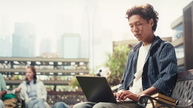 Portrait shows a smiling student relaxing on a wooden campus bench, typing notes on a laptop with stacked books nearby during an independent study session.