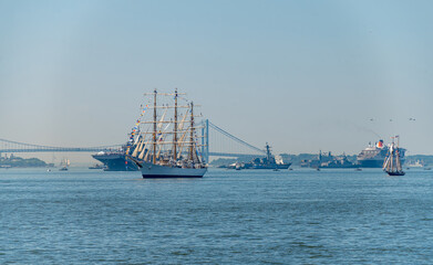 Tall ship leads Independence Day fleet near Verrazzano Narrows Bridge. Rigged sailing vessel moves through blue harbor water among naval ships, cruise liners and smaller sailboats. © white78