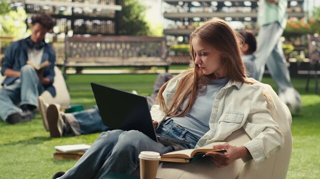 Student sits on a beanbag holding an open book and a laptop, looks up during independent outdoor study and keeps a notebook nearby on a university campus with peers in the background.