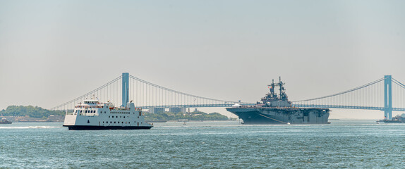 USS Kearsarge and passenger ferry cross Verrazzano Narrows Bridge. Amphibious assault ship and white ferry occupy blue harbor water with suspension bridge behind. © white78