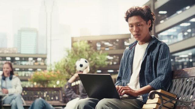 Focused international student checks notes on a laptop from a campus bench, with stacked books and a coffee cup nearby in a quiet independent outdoor learning session for exam preparation.