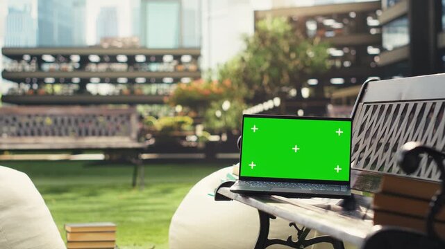 Green screen mockup on a laptop rests on a modern campus bench outdoors with blurred buildings behind, providing an editable display for education content and screen replacement.