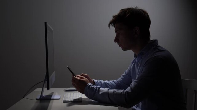 Man ignoring his computer screen and focusing intently on his smartphone, working late or checking messages in a dimly lit office environment, depicting themes of digital distraction and overwork