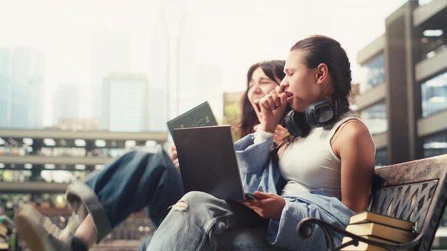 Student friends laugh and joke together on a campus bench beside an open laptop during a relaxed peer tutoring and academic study session.
