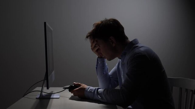 Man covering his face with his hand, sitting in front of a computer screen, holding a game controller and experiencing frustration and disappointment after losing a video game in a dimly lit room