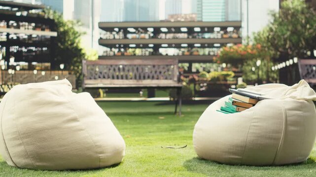 Multiple stacked books and study materials rest on a beanbag outdoors at a modern university campus, forming an empty academic learning setup with literature, research tools and a notebook.