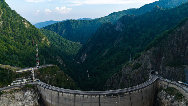 Aerial view of the massive concrete Vidraru Dam nestled in a deep mountain valley surrounded by dense green forests and winding roads in Arefu, Arges, Romania.