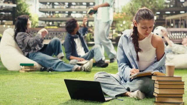 Woman student sits on a university campus lawn and writes notes from an open book, with stacked books and a backpack nearby during an outdoor learning session.