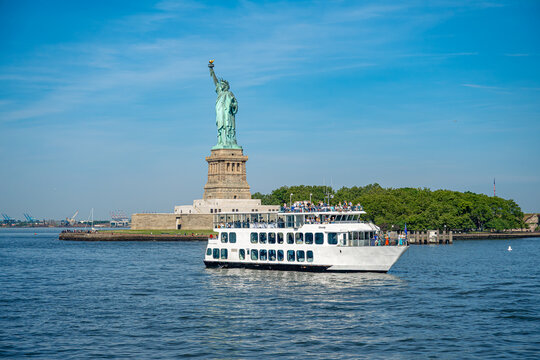 New York, USA, 4 Jule 2026 : Tourist ferry cruising past Statue of Liberty in New York. White double deck ship sails near Liberty Island on calm water under sunny sky.