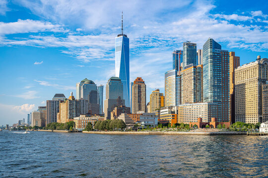 Financial district skyline along Hudson River in New York City. Tall office buildings and green waterfront park stretch under blue sky.