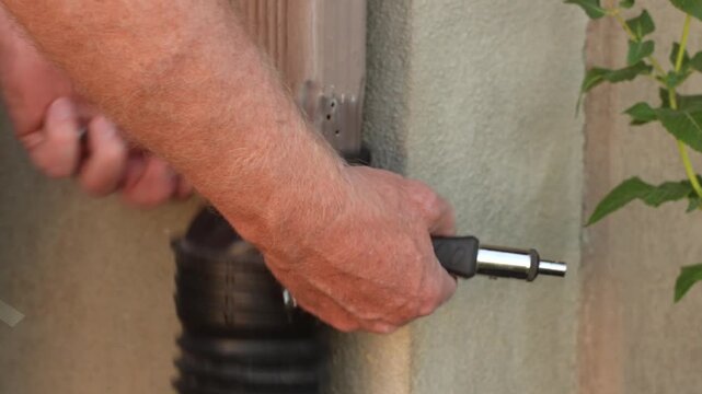 Close-up of a homeowner using a hand tool to fasten a black downspout adapter to an exterior rain gutter. 